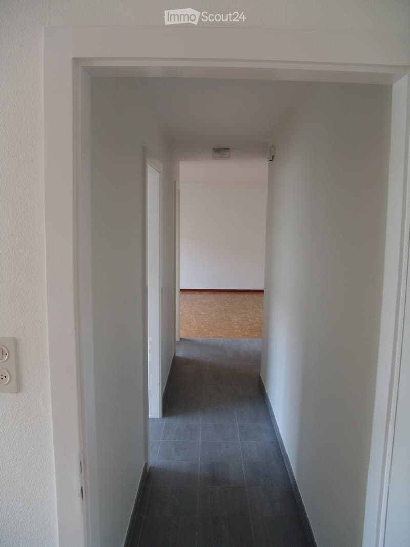Empty hallway with tiled floor and white walls, leading to another room with wooden flooring visible at the end.