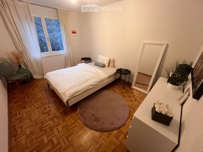 Single bed, night table, round carpet, mirror, shelf with flowers and plant, gray chair, white curtains, parquet flooring