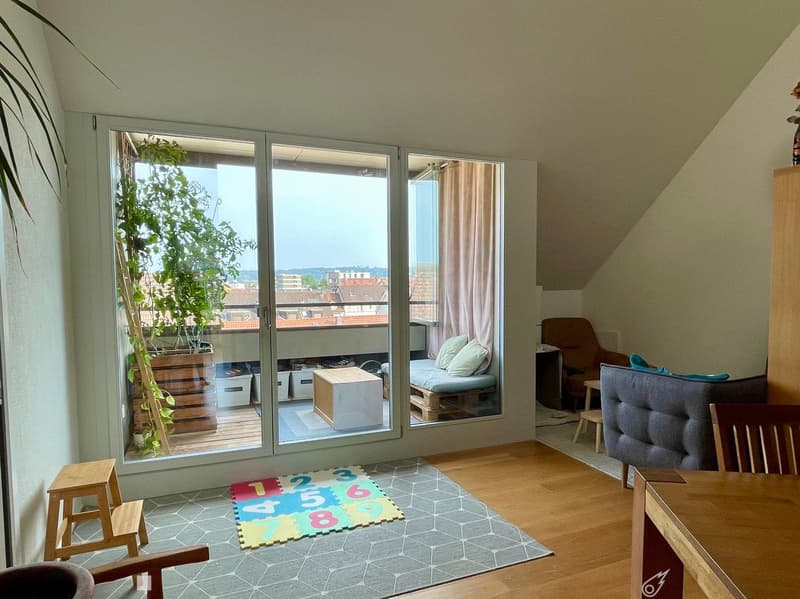 Bright living room with large windows and a balcony, featuring a sofa, chairs, a small table, and a colorful play mat on the floor.