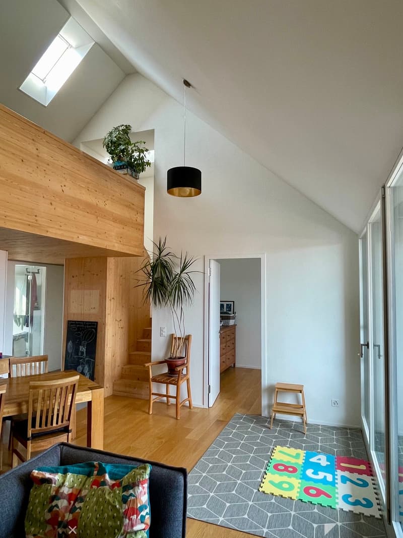 Bright, open living space with wooden floors, a grey sofa with colorful pillows, a wooden dining table with chairs, a hanging lamp, a potted plant, and a colorful foam mat with numbers on the floor.