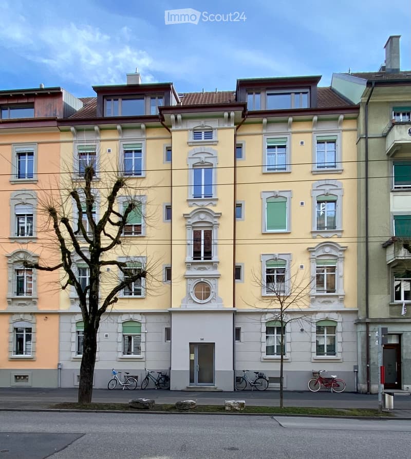 Yellow building with a chimney, yellow and gray facade, multiple windows, bicycles parked in front, balcony in the top right corner, tree in front.