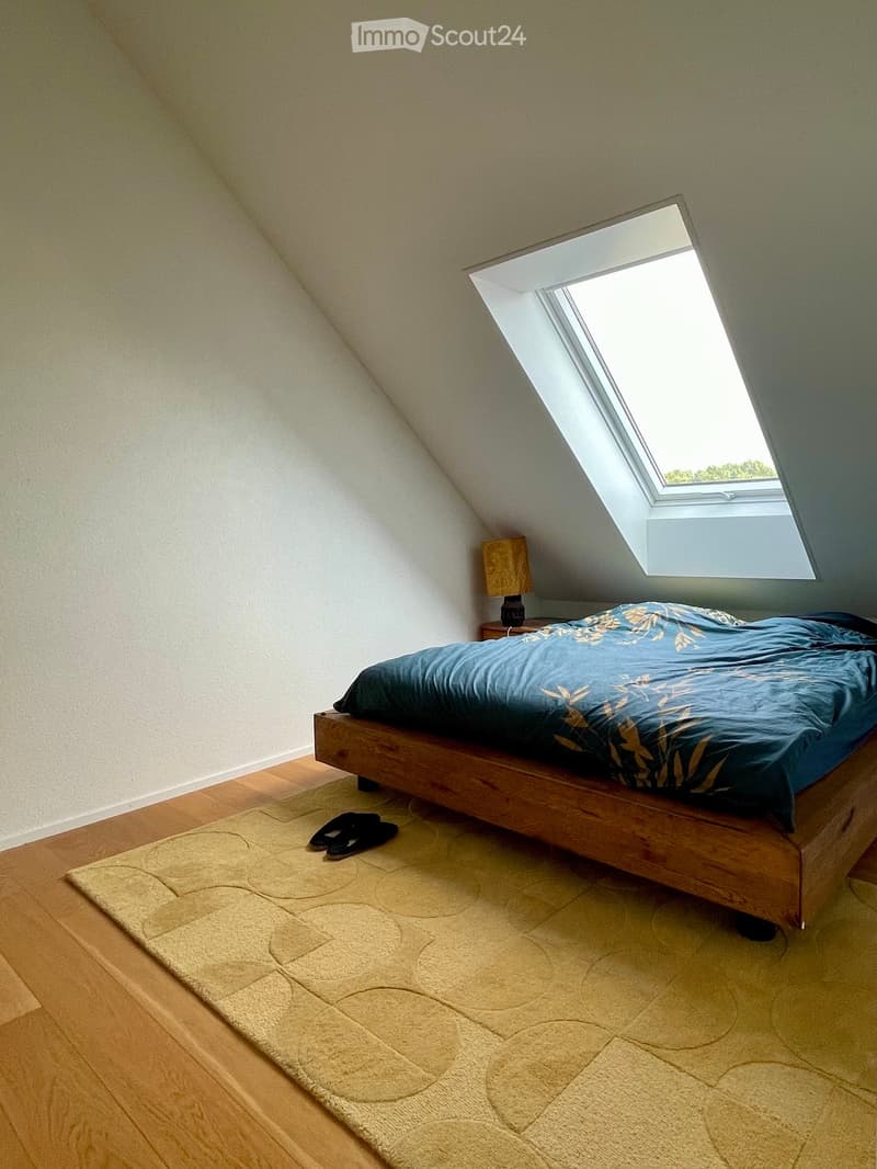 Cozy attic bedroom with wooden floor, slanted ceiling, skylight window, bed with blue duvet, nightstand with lamp, and a small rug.