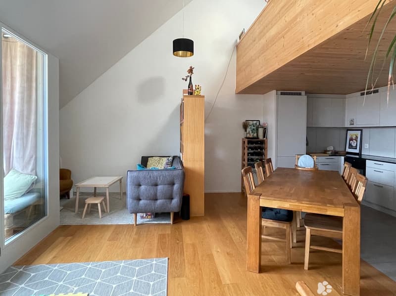 Bright, open-plan living and dining area with wooden flooring, large windows, a grey sofa, a small coffee table, a dining table with chairs, a wine rack, and a modern kitchen area with cabinets and appliances.