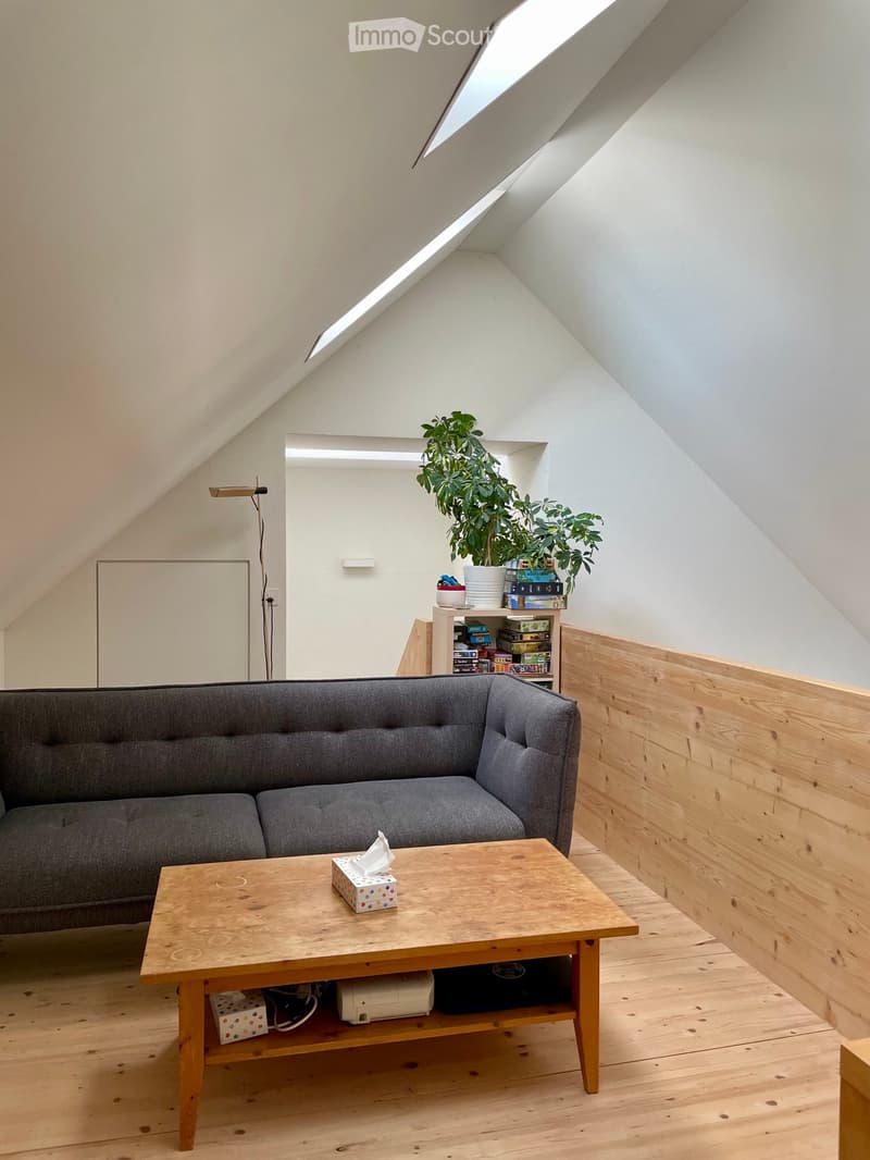A cozy attic living room with wooden floors, a grey sofa, a wooden coffee table, a potted plant, and a shelf with books and decorative items.