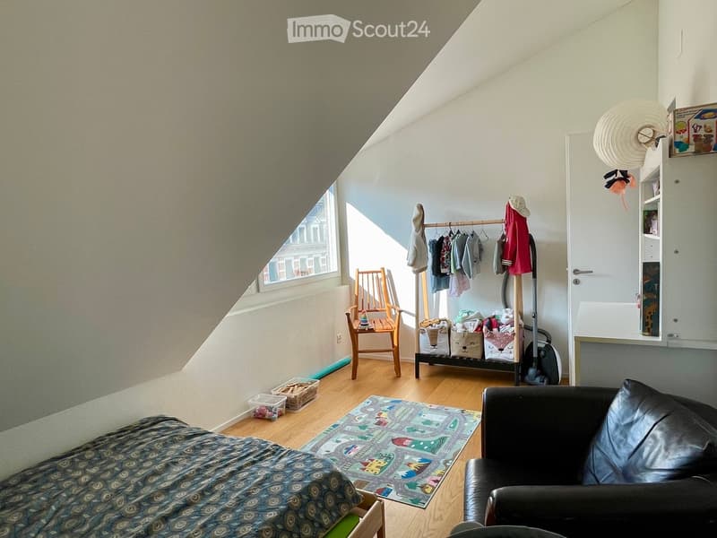 Cozy attic bedroom with slanted ceilings, wooden flooring, and large window. Features a bed, sofa, wooden chair, coat rack, and play mat.