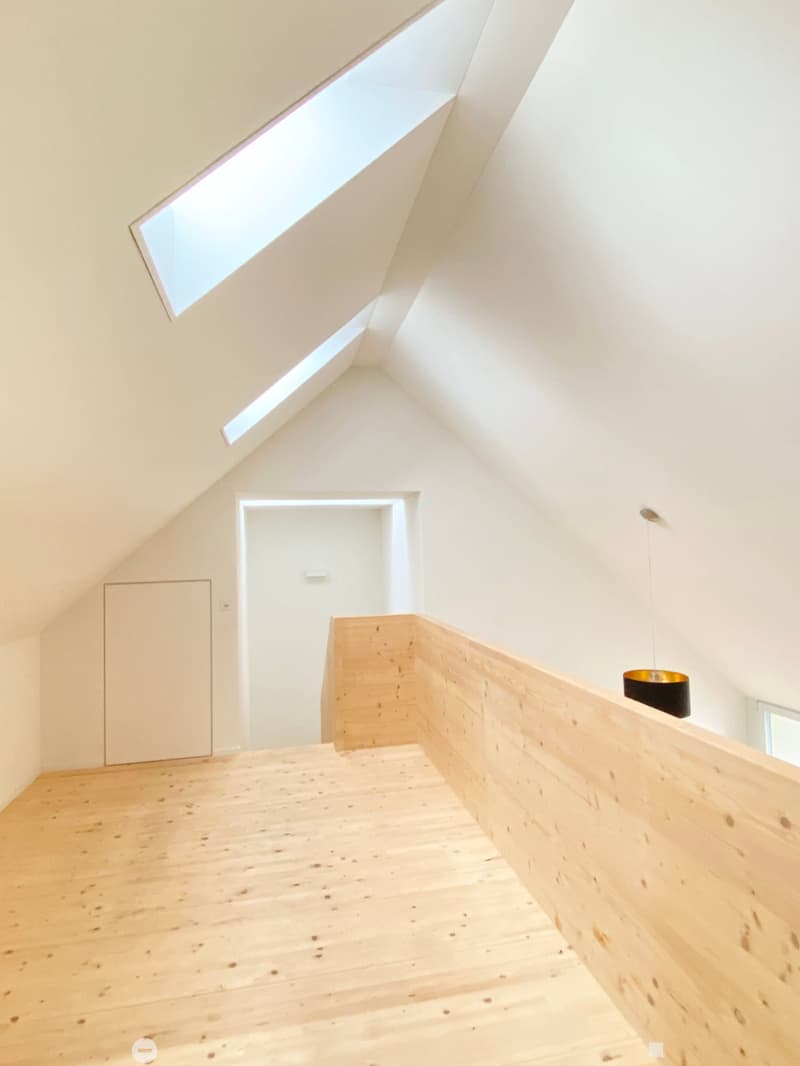 Bright, minimalist hallway with wooden flooring, white walls, and a skylight at the top. Features a wooden railing and a single hanging light fixture.