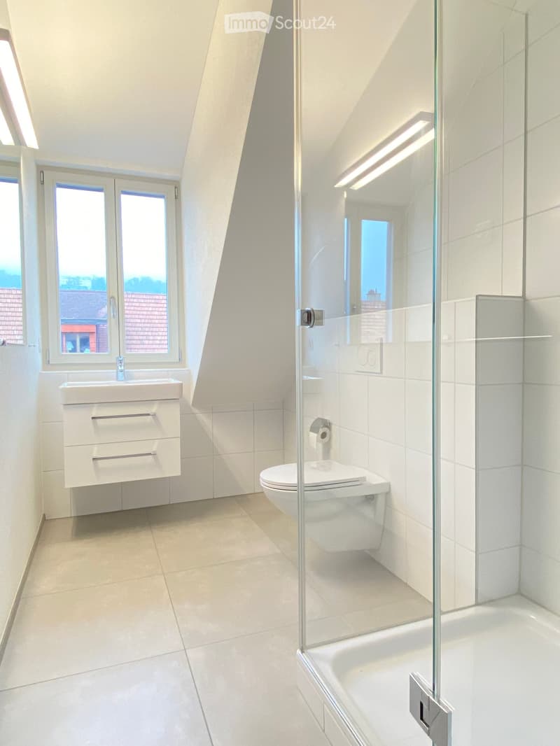 Modern bathroom with white tiles, a sink, toilet, bathtub, and a glass-enclosed shower stall. Large windows provide natural light.