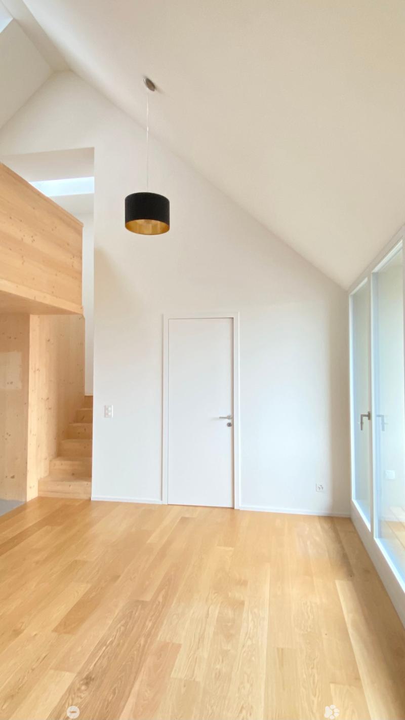 Empty room with light wooden flooring, white walls, and a hanging pendant light. Features a staircase on the left and a closed door in the center.