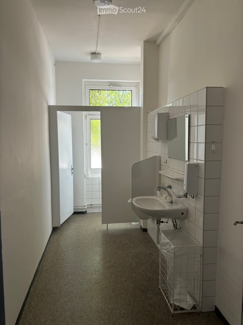 Clean, well-lit hallway with carpeted floor, white walls, and a window at the far end. Features a sink area with a mirror, soap dispenser, and a trash bin.
