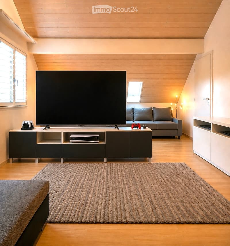 Modern living room with wooden flooring, grey sofa, large flat-screen TV on a black and white entertainment unit, gaming console and controllers, floor lamp, and a woven area rug.