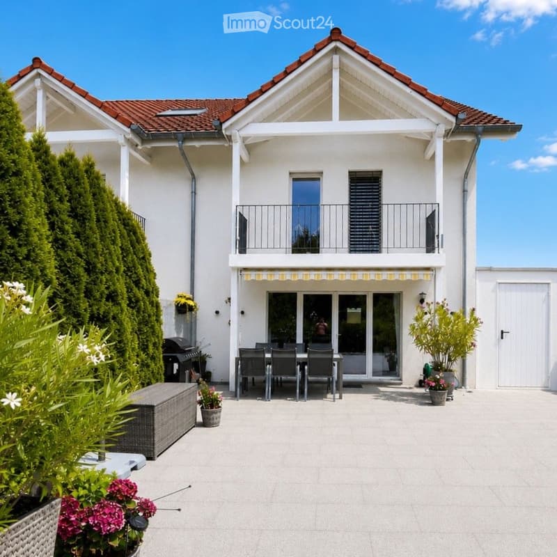 Modern two-story house with a balcony and a patio area featuring a dining table, chairs, a bench, and potted plants.