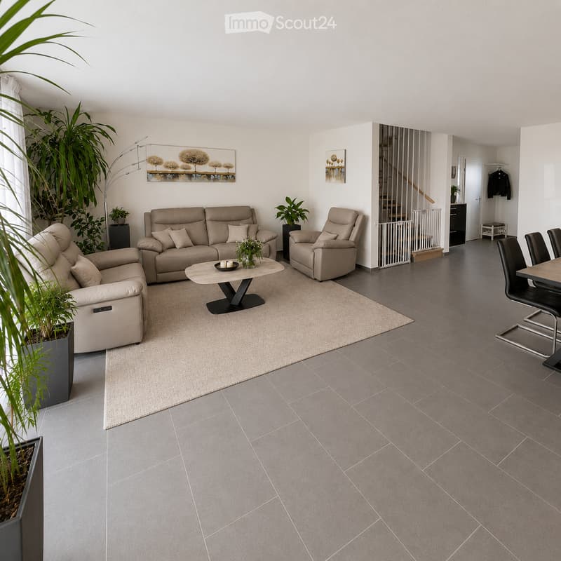 Modern living room with grey tiled floor, beige sofa, armchair, coffee table, dining area with chairs and table, large potted plant, artwork on walls.