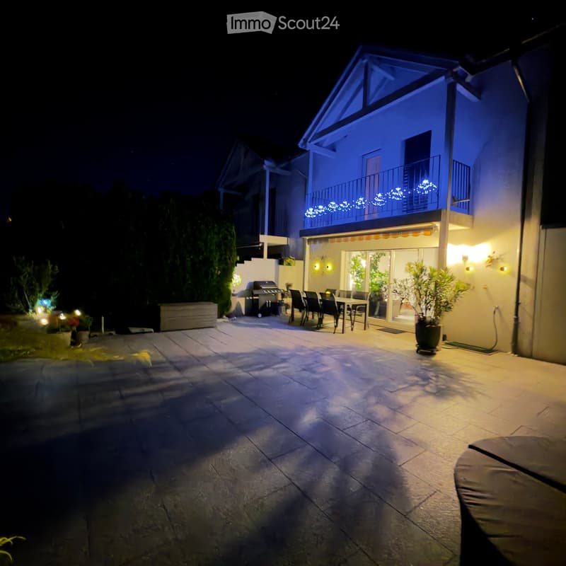 Well-lit outdoor patio with stone tiles, a dining table with chairs, a grill, potted plants, and string lights on the balcony above.