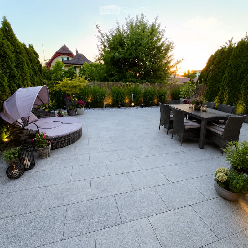 A well-maintained patio with a grey stone tile floor, featuring a round wicker sofa with cushions, a rectangular dining table with four chairs, and several potted plants and candle holders.