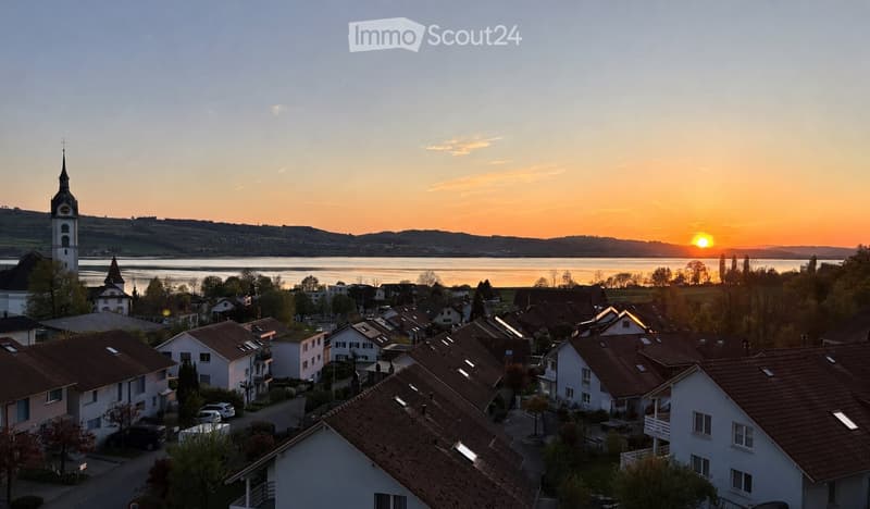 A village scene at sunset with houses, a church tower, and a body of water in the background.