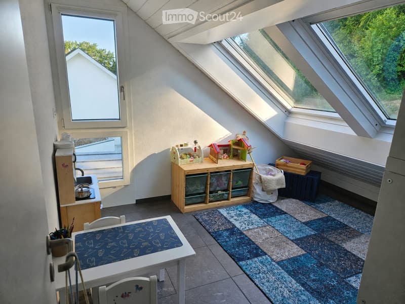 A cozy, well-lit attic room with skylights, featuring a small kitchenette, a play table, a toy box, and a colorful rug.