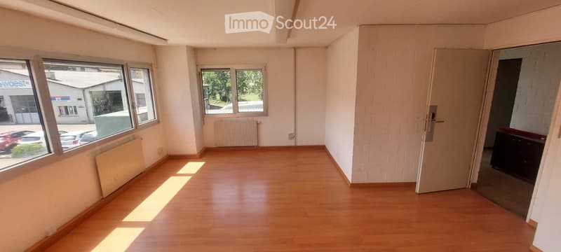 Empty room with wooden flooring, white walls, and large windows allowing natural light. Radiators are visible along the walls, and there is a closed door leading to another space.