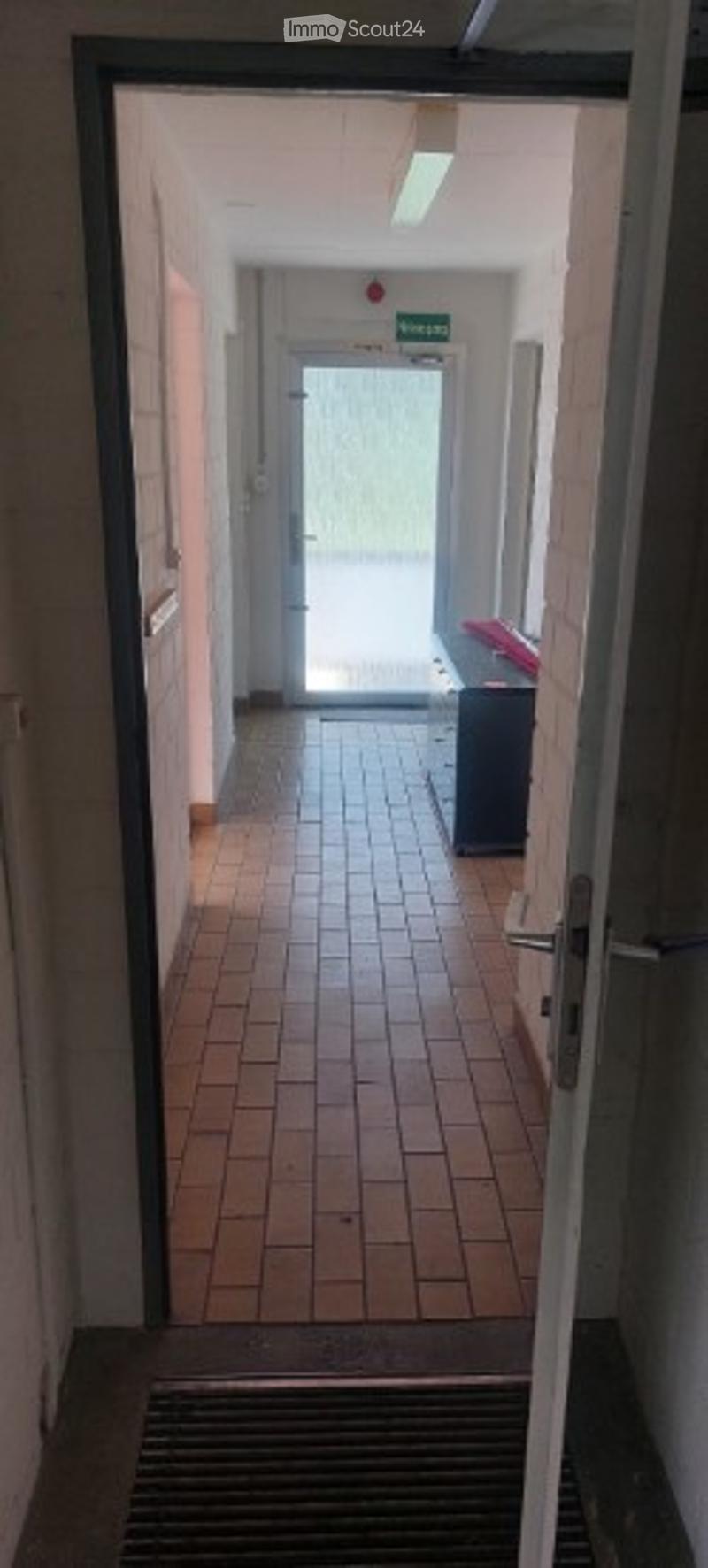 A narrow hallway with tiled flooring, leading to a door at the end. The walls are painted in a light color, and there is a small cabinet visible on the right side.