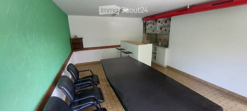 A small, furnished room with a black table, several black chairs, a small white cabinet, and a small wooden shelf. The walls are painted green and white, and the floor is tiled.
