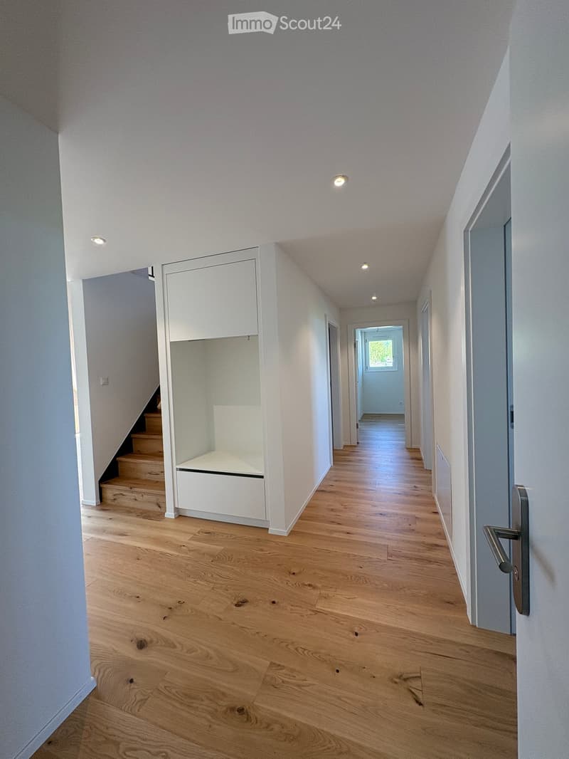 Clean, modern hallway with light wooden flooring, white walls, recessed ceiling lights, and a built-in storage unit with a bench seat. A staircase leads upwards, and a doorway at the end opens to another room.