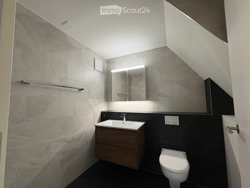 Modern bathroom with a white sink, wooden cabinet, toilet, and large mirror with built-in lighting. Grey marble tiles on walls and dark tiles on floor.