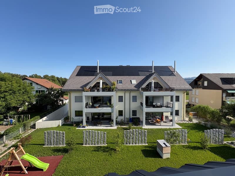 Modern two-story house with white exterior, solar panels on the roof, multiple balconies, and a well-maintained lawn with a small play area featuring a slide and wooden play structure.
