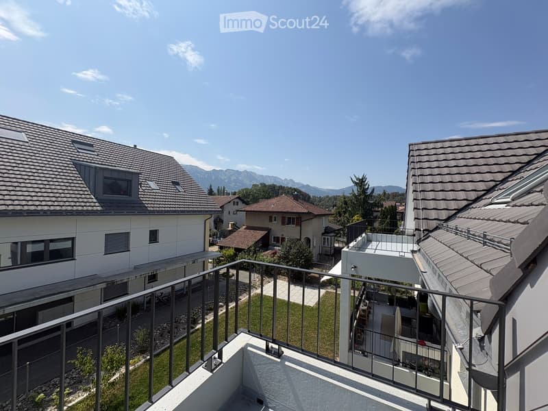 Balcony overlooking a residential area with houses, a lawn, and distant mountains under a clear blue sky.