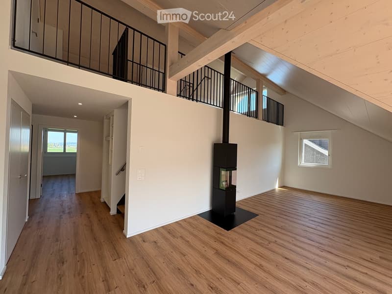 Open-plan living space with wooden flooring, white walls, and a modern black wood-burning stove. Features include exposed wooden beams, a staircase leading to an upper level, and large windows offering natural light.