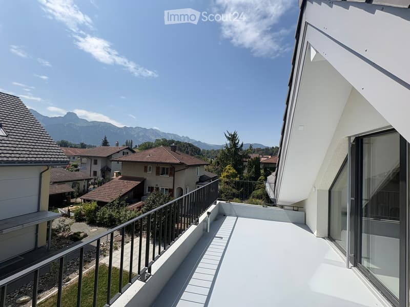 Balcony with white floor and metal railing, overlooking a residential area with houses, trees, and distant mountains.