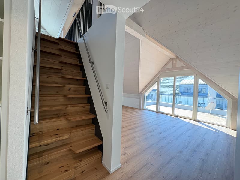 Modern wooden staircase with metal railing leading to a bright, open loft area with large windows and wooden flooring.