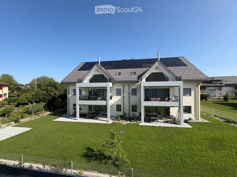 Two-story modern house with solar panels on the roof, large windows, balconies, and a well-maintained lawn.