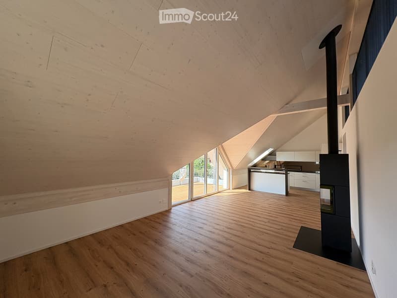 Open-concept living space with wooden floors, exposed wooden ceiling beams, and a modern kitchen area featuring white cabinets and a central island. A wood stove is positioned near the center.