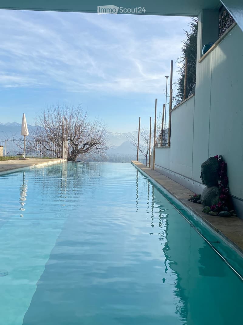 A large swimming pool with clear blue water, surrounded by a white wall and wooden poles, with a scenic mountain view in the background.