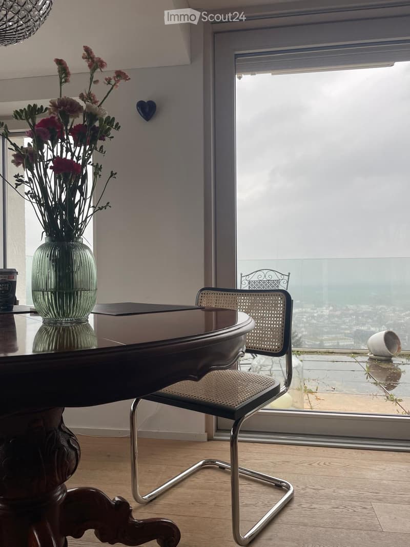 A modern, furnished room with a wooden floor, a large window offering a scenic view, a dark wooden table with a vase of flowers, and a sleek chair with a woven seat.