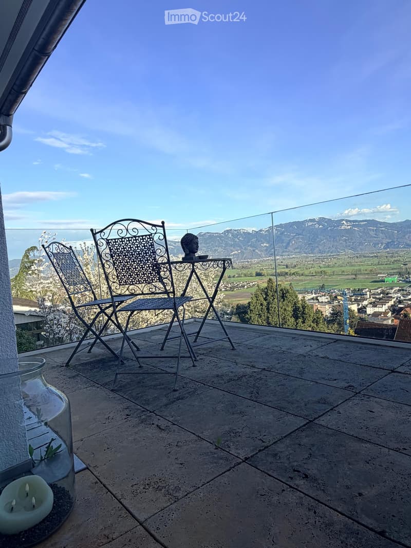 Balcony with a view of mountains and cityscape, featuring two metal chairs, a small table, and a candle in a glass container.