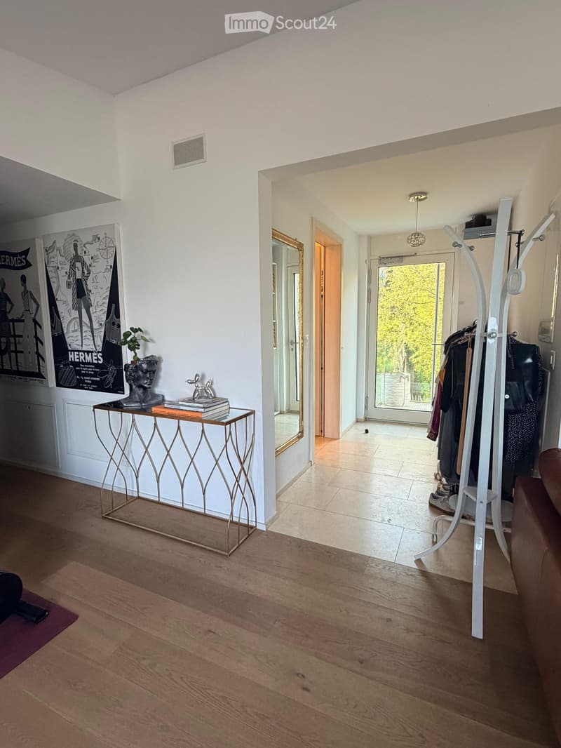 A modern, well-lit hallway with wooden flooring, a small table with books and a statue, a coat rack with clothing, and a large mirror on the wall.