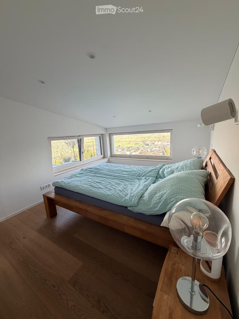 Modern bedroom with a wooden bed, light blue bedding, and a sleek glass lamp on a nightstand. Large windows offer a scenic view.