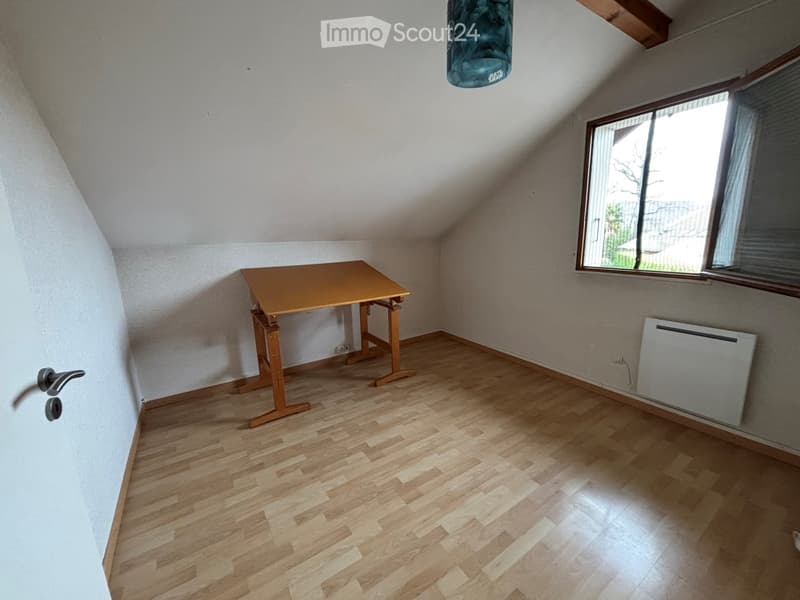 A small, unfurnished room with wooden flooring, a single table, and a window with blinds. The room has white walls and a slanted ceiling typical of an attic space.