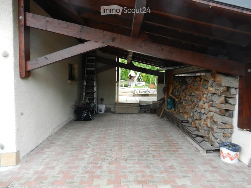 Brick-paved outdoor area with wooden ceiling beams, stone woodpile, ladder, and various stored items.