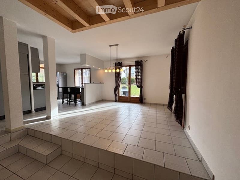 Empty room with tiled floor, wooden ceiling beams, and large windows with dark curtains. There is a small kitchen area visible in the background.