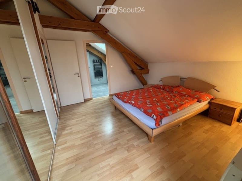 Cozy attic bedroom with wooden beams, a double bed with a colorful duvet, a nightstand, and exposed wooden beams on the ceiling.