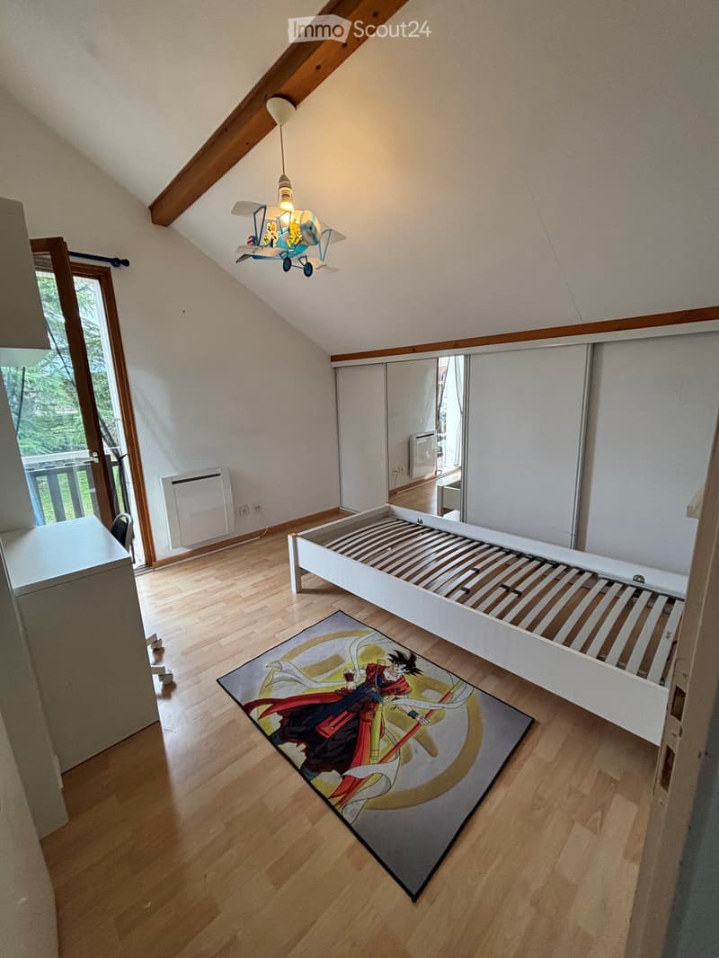 Spacious attic bedroom with wooden floors, large window, built-in wardrobe, single bed, colorful anime-themed rug, and a hanging airplane toy light fixture.