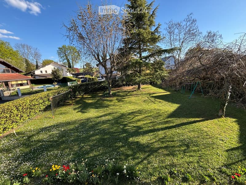 Well-maintained garden with green grass, colorful flowers, trees, and a swing set in the background.