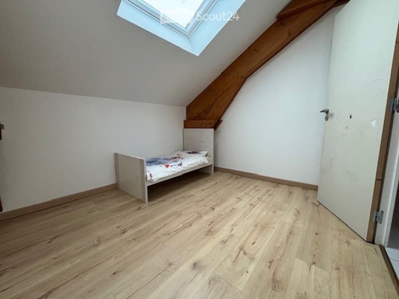 Small bedroom with wooden flooring, white walls, and a single bed against the wall. The room has a slanted ceiling with exposed wooden beams and a skylight window.