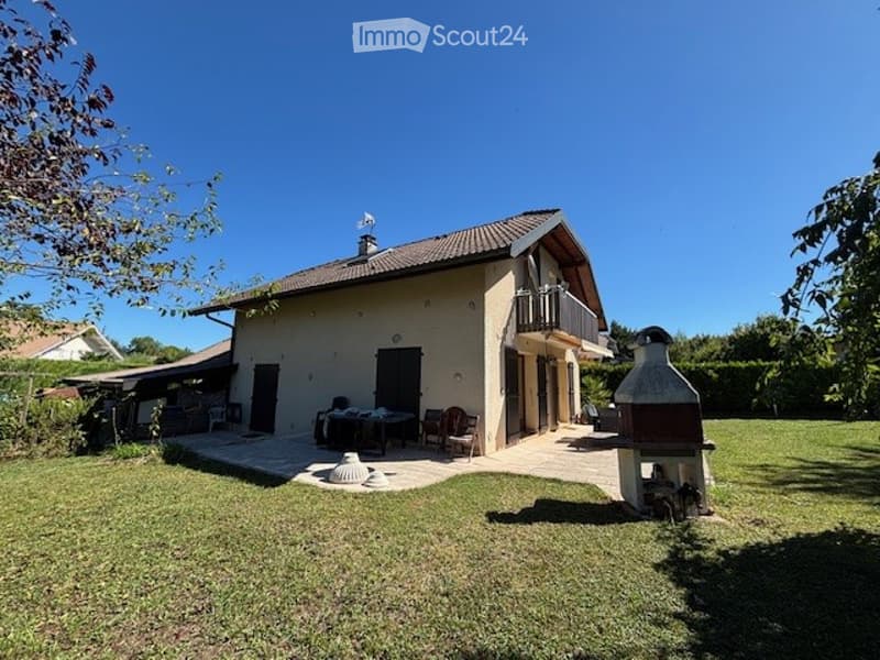 Single-story house with beige exterior, tiled roof, and a small balcony. The surrounding area includes a well-maintained lawn, outdoor furniture, and a barbecue area.
