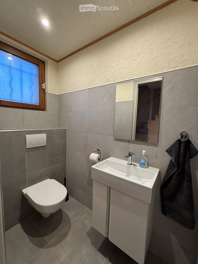 Modern bathroom with grey tiled walls and floor, white toilet, sink, and mirror, towel hanging on the wall, and a small window with frosted glass.