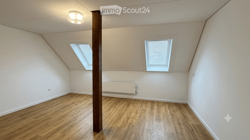 Empty attic room with wooden flooring, white walls, and two skylight windows. Features a central support beam and a radiator on one wall.