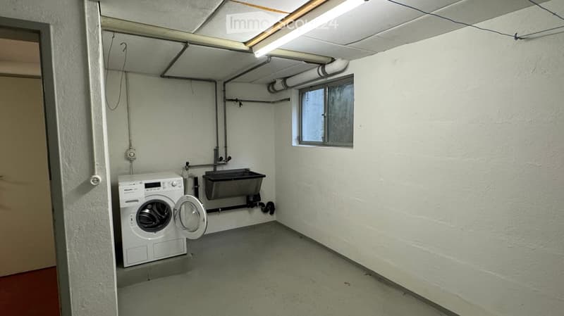 Utility room with a washing machine, sink, and exposed pipes. The room has white walls, a concrete floor, and a small window.