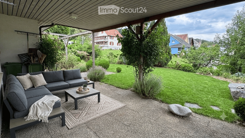 Outdoor patio with a grey sectional sofa, coffee table, small side table, and a patterned rug on a gravel surface, surrounded by a well-maintained lawn and garden.