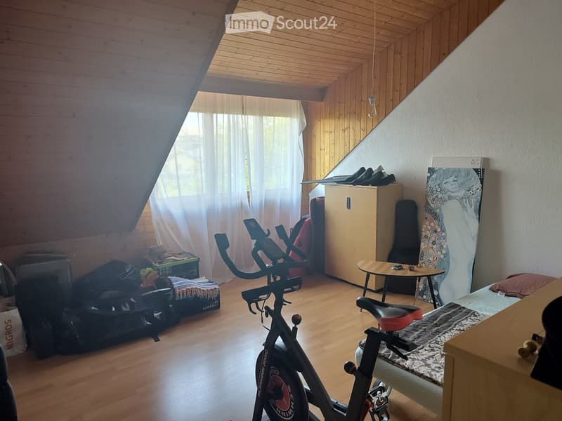 A cozy attic room with wooden walls and ceiling, featuring an exercise bike, a bed with patterned bedding, a small table with a chair, a cabinet, and a guitar leaning against the wall.